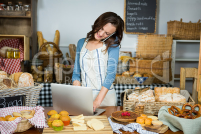 Smiling staff using laptop while talking on mobile phone at bakery counter