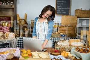 Smiling staff using laptop while talking on mobile phone at bakery counter