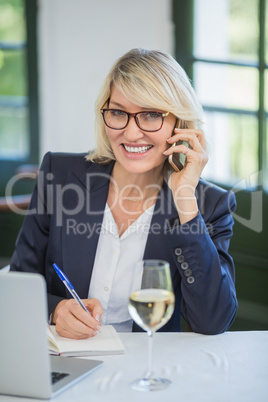 Businesswoman talking on mobile phone while writing on diary