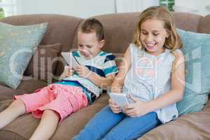 Smiling sister and brother sitting on couch using mobile phone in living room