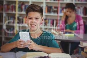 Smiling schoolboy using mobile phone in library at school