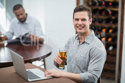 Handsome man holding beer glass while using laptop