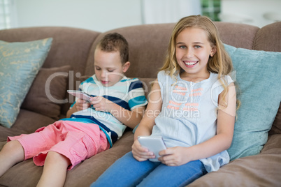 Smiling sister and brother sitting on couch using mobile phone in living room