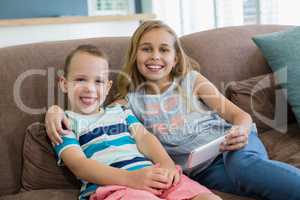 Smiling sister and brother sitting on couch using mobile phone in living room at home