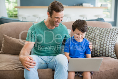 Smiling father and son sitting on sofa using laptop in living room