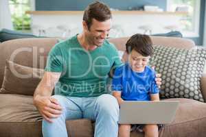 Smiling father and son sitting on sofa using laptop in living room