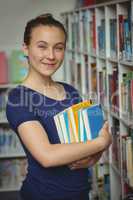 Portrait of schoolgirl standing with stack of books in library
