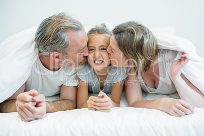 Two parents lying on the bed while kissing their daughter under a blanket