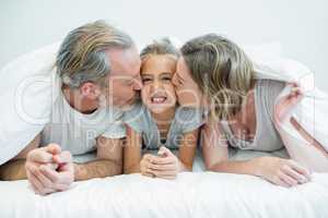 Two parents lying on the bed while kissing their daughter under a blanket