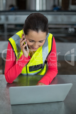 Female factory worker using laptop while talking on mobile phone