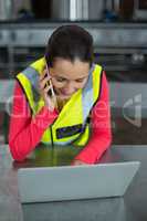Female factory worker using laptop while talking on mobile phone