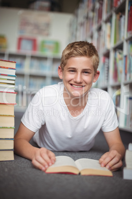 Portrait of happy schoolboy lying on floor with books in library