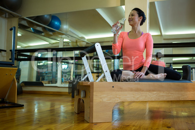 Happy woman sitting on reformer and drinking water after workout