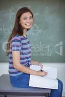 Portrait of schoolgirl sitting on bench and writing on book in classroom