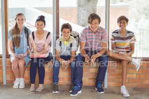Portrait of happy students sitting on window sill and using mobile phone in corridor