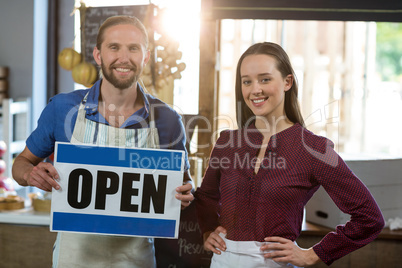 Portrait of smiling bakery staff standing with open sign board at counter