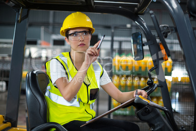 Female factory worker sitting on forklift and writing in clipboard at factory