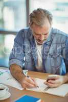 Male business executive writing in organizer while using mobile phone