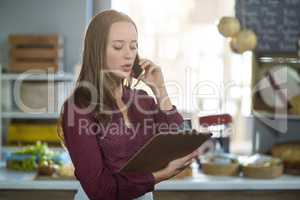 Female staff checking clipboard while talking on mobile phone at counter