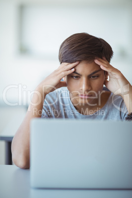 Tensed schoolboy looking at laptop in classroom