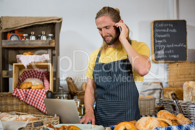 Smiling male staff talking on mobile phone while working on laptop at counter