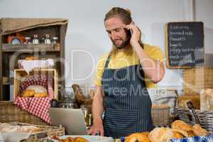 Smiling male staff talking on mobile phone while working on laptop at counter