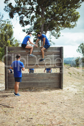 Female trainer assisting fit man to climb over wooden wall during obstacle course