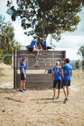 Female trainer assisting fit woman to climb over wooden wall during obstacle course