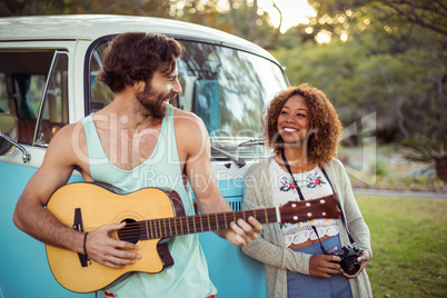 Man playing guitar near campervan while woman standing beside him