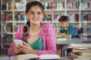 Smiling schoolgirl using mobile phone in library at school