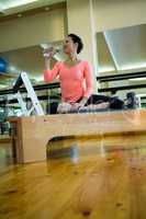 Happy woman sitting on reformer and drinking water after workout
