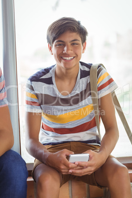 Portrait of happy schoolboy sitting on window sill and using mobile phone in corridor