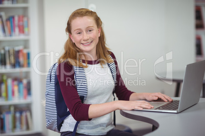 Portrait of happy schoolgirl with schoolbag using laptop in library