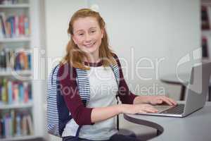 Portrait of happy schoolgirl with schoolbag using laptop in library