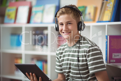 Portrait of happy schoolboy listening music while using digital tablet in library