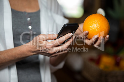 Mid section of female costumer holding orange while using mobile phone