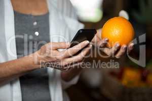 Mid section of female costumer holding orange while using mobile phone