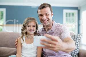 Smiling father and daughter sitting on sofa and taking selfie in living room