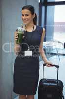 Beautiful female executive standing with luggage while having juice in corridor