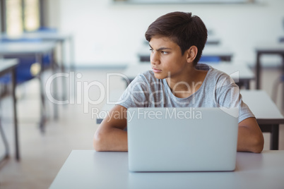 Thoughtful schoolboy sitting with laptop in classroom