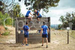 Female trainer assisting fit woman to climb over wooden wall during obstacle course