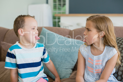 Sister and brother stick out tongues to each other on couch in living room