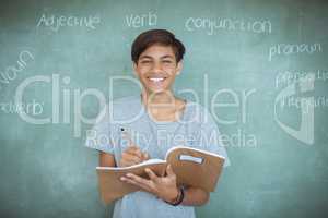 Portrait of schoolboy writing in notebook against chalkboard in classroom