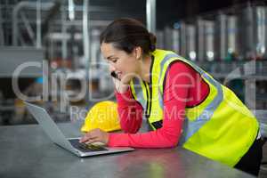 Female factory worker using laptop while talking on mobile phone
