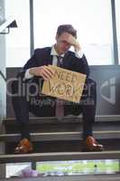 Depressed businessman sitting on stairs holding cardboard sheet with text need work