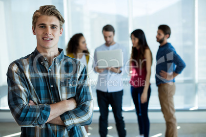 Man standing with arms crossed in office while colleague discussing in background Man standing with arms crossed in office while colleague discussing in background