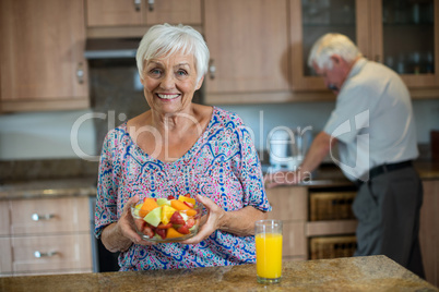 Senior woman holding bowl of fruit while man working in kitchen