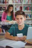 Smiling schoolboy using mobile phone in library at school