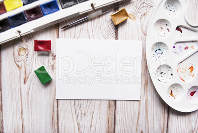 Artist workspace with watercolor, brush, palette, white paper on a wooden background.