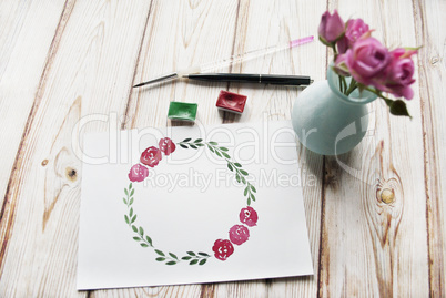 Artist workspace with watercolor, brush, bouquet of roses, hand drawn floral wreath frame on a wooden background.